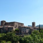Vista panorámica del pueblo medieval de Santa Pau, en Cataluña, España. La iglesia de Santa María y el Castillo de Santa Pau se ubican en primer plano, rodeados de montañas y bosques.